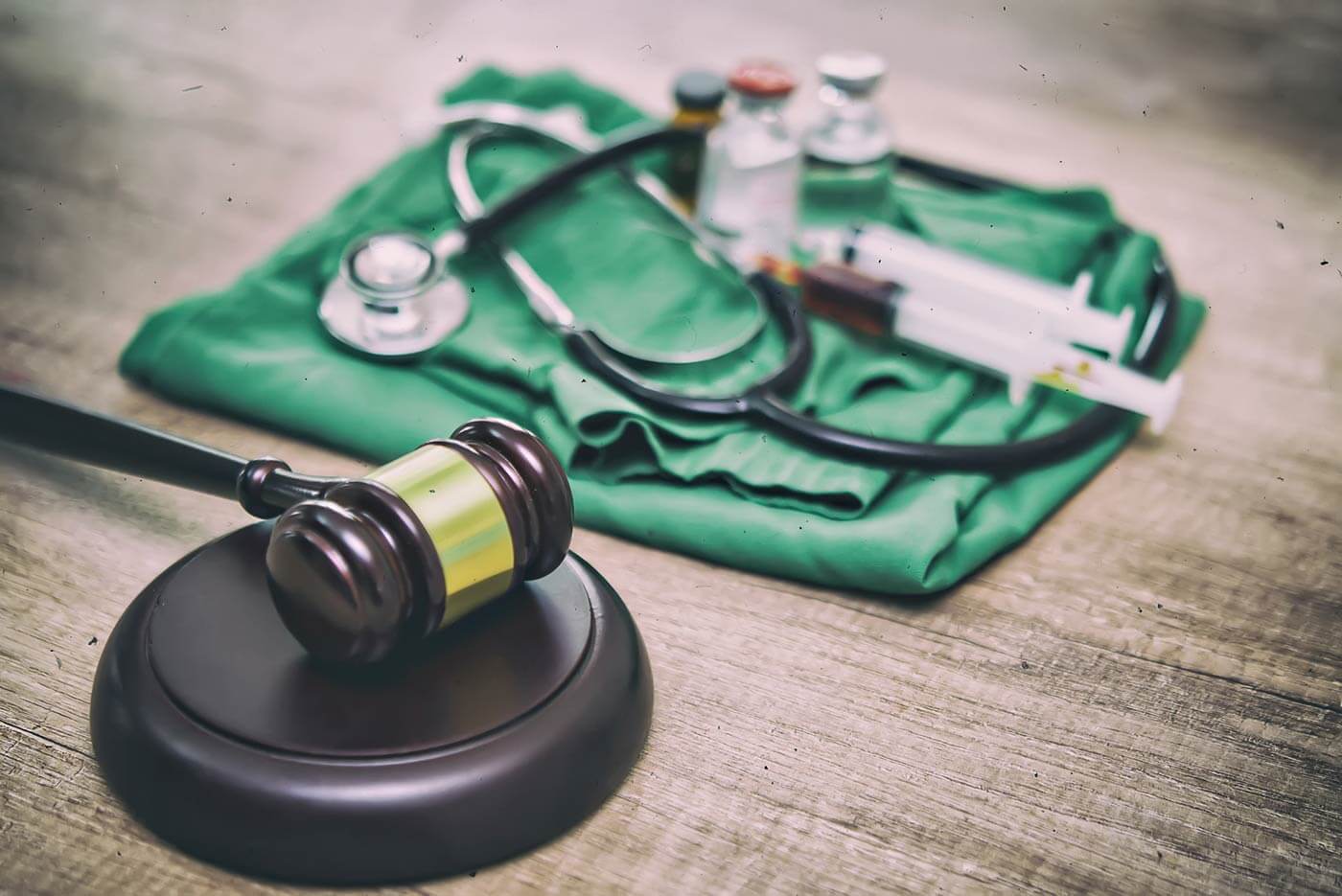 Folded surgical scrubs, stethoscope, medicine vials,  and a judge's gavel on a distressed wooden surface.